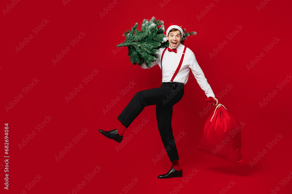 Poster Full body merry young cool man wearing white shirt Santa hat posing hold show bag with present gifts Christmas tree isolated on plain red background studio. Happy New Year celebration holiday concept.