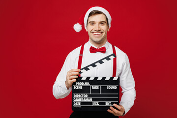 Merry young man wears white shirt Santa hat posing hold classic black film making clapperboard isolated on plain red background studio portrait. Happy New Year Christmas celebration holiday concept.