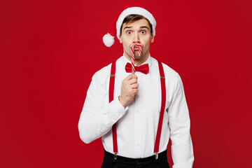 Merry young surprised man wear white shirt Santa hat posing hold in hand candy cane on lips look...