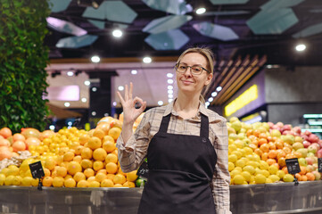 Young fun seller cashier woman wearing uniform show ok okay working at supermaket store grocery shop standing near exotic fruit products selling inside hypermarket. Purchasing food gastronomy concept.