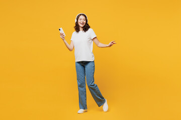 Full body young fun woman she wear white blank t-shirt casual clothes listen to music in headphones use mobile cell phone isolated on plain yellow orange background studio portrait. Lifestyle concept.