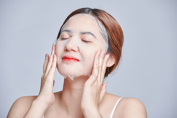 Happy young removing from face skincare hydrating sheet mask with collagen, hyaluronic acid for glowing skin. daily self care routine. Photo of attractive woman with perfect skin on grey background.
