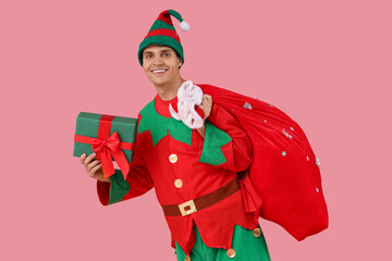 Handsome young man in elf's costume with Santa bag and gift box on pink background