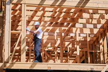 Naklejka na ściany i meble Carpenter building wooden-framed house. Bearded man donning a protective jumpsuit and helmet, inspects the walls for levelness using a spirit level. Concept of environmentally friendly construction.