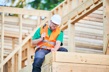 Carpenter constructing wooden frame house. Bearded man wearing glasses hammering nails into...