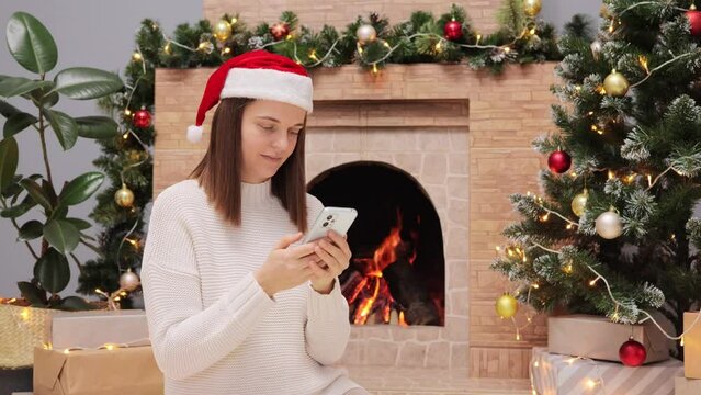 Dark haired adult woman wearing warm sweater and Santa Claus hat sitting in living room near fireplace and Christmas tree holding cell phone reading posts in network looking at festive photos.