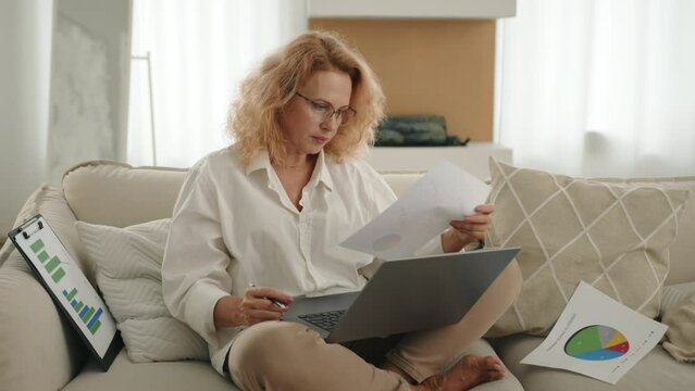 Puzzled middle-aged blonde woman in glasses with short curly hair looking attentively into laptop, making notes with pencil on shit of paper with pie chart. High quality 4k footage