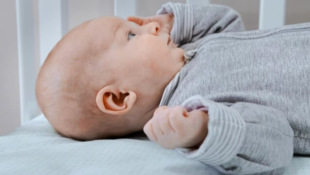 Alone toddler is lying calmly on his back in the crib, day. Newborn infant has insomnia. Suckling wearing little onesie. Baby does not sleep during bedtime. Babe dressed in boilersuit, coverall