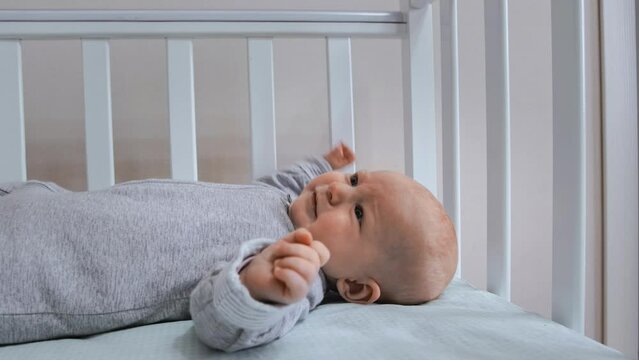 Infant is lying on his back in the crib, newborn crying and acting up, day. Naughty baby has insomnia. Alone toddler does not sleep during bedtime. Suckling wearing little onesie boilersuit, coverall