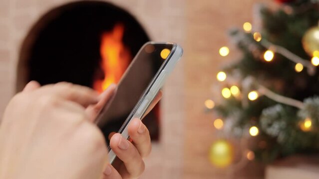 Female hands holding phone with blank screen over decorated Christmas tree fireplace scrolling online browsing internet chatting with friends winter shopping.