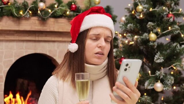 Sick woman using mobile phone for making video call holding glass of champagne sitting near Christmas tree and fireplace at home communicating online.