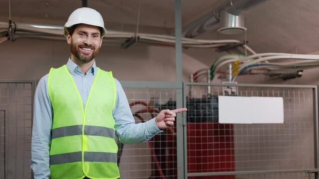 Smiling experienced professional electrician points to white signboard for advertising at electrical network station Male engineer making hand gesture for text insertion copy space or template layout.
