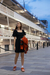 Young girl in black dress with red jacket and headphones in hands is standing on pier against passenger liner and beautiful evening sky. She is waiting for someone. Travelling, vacation, tourism