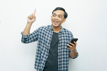 Naklejka premium Portrait Indonesian Asian young man smiling while looking at smartphone and index finger pointing up. Isolated white background