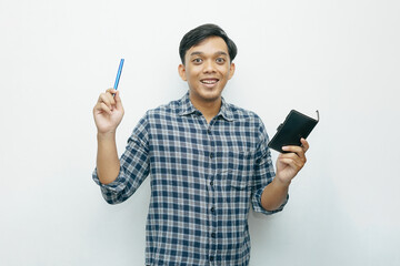 Portrait of young Asian Indonesian man smiling when holding small notebook with ballpoint pen with isolated white background