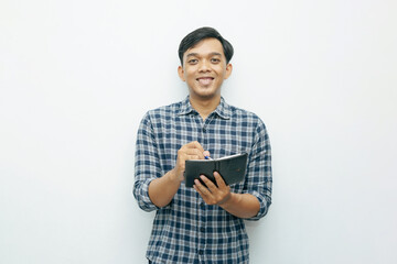 Portrait of young Asian Indonesian man smiling when writing on small notebook with ballpoint pen...
