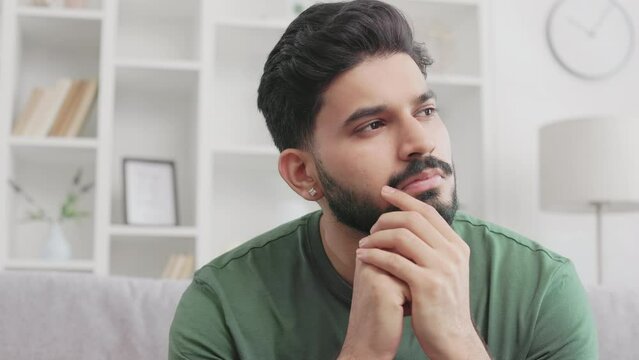 Close up portrait of pensive hindu guy scratching bearded chin while lost in thoughts at home. Bearded young man in green t-shirt sitting on cozy sofa and looking aside seriously.