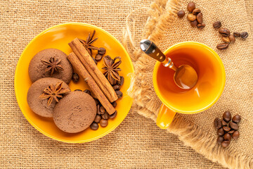 Chocolate cookies, cinnamon, star anise, coffee beans on a ceramic saucer with a cup on a jute cloth, macro, top view.