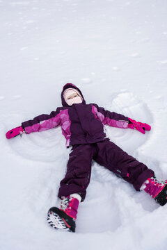 A child in a purple snowsuit lies spread-eagled in the snow, creating a perfect snow angel, her face alight with joy.