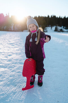 A girl in a knit hat and winter coat holds a red snow sled, giving a thumbs up on a snowy slope.