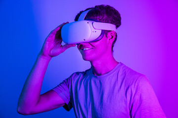 Fototapeta premium Smiling young guy with VR headset exploring virtual reality isolated on blue and pink neon background