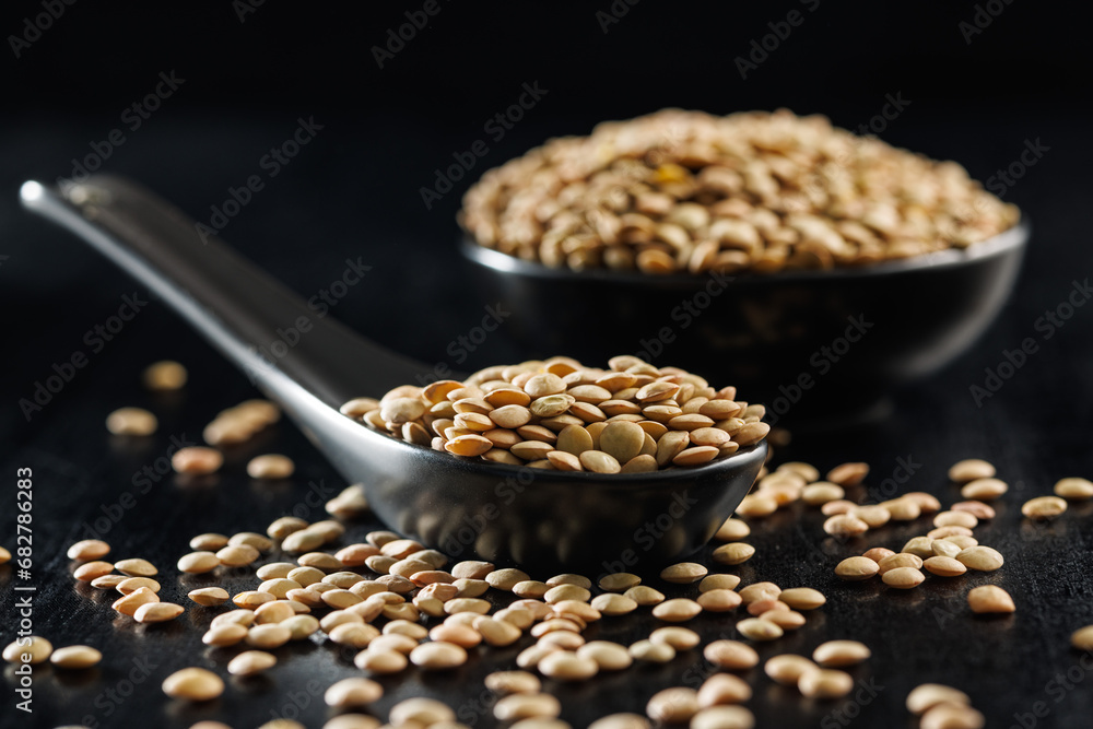 Sticker uncooked lentil legumes in ceramic spoon on black table.