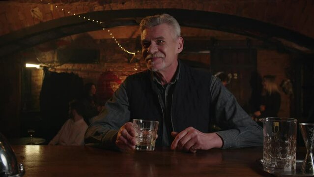 Man chatting with barman while drinking his armenian cognac. Positive elderly male discuss to bartender in bar. Free time spending summer evenings at retro pub. Mature alcoholic drinks vodka