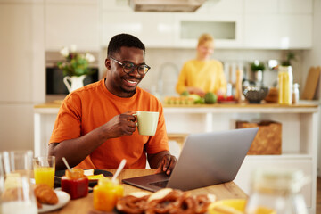 African American young man using laptop during breakfast at table in the kitchen with his girlfriend cooking in background