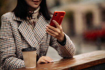 A woman in an autumn coat and scarf holds a phone in her hands in a cafe on the street, drinks takeaway coffee. Close-up