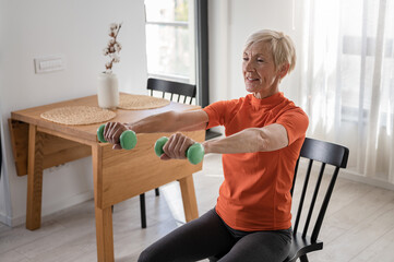 Smiling beautiful senior woman health instructor doing chair exercises with dumbbells