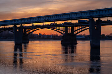 Beautiful view of the bridge over which cars drive at sunset. A river flows under the bridge, reflecting the sunset rays. Bridge in the city of Novosibirsk, Russia