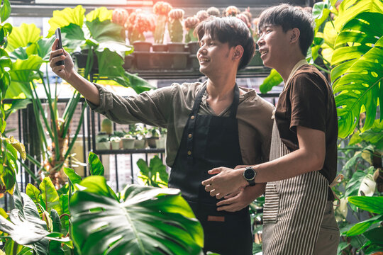 Two Asian male plant shop owners in aprons holding smartphones send pictures of plants to customers. People order plants. Success in the plant shop. Small business owner ideas