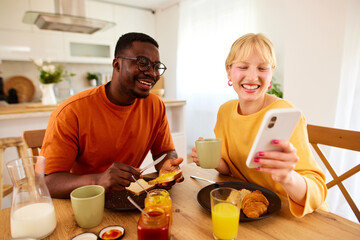 Happy multiracial couple using mobile phone together over breakfast at home