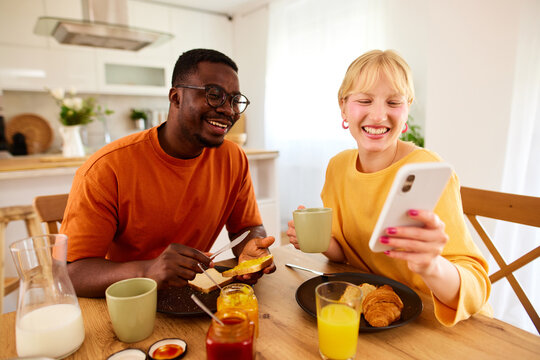 Happy Multiracial Couple Using Mobile Phone Together Over Breakfast At Home