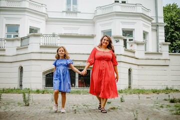 Naklejka premium Happy mother inred dress and daughter in blue walk hand in hand towards camera.Harmonious family. 