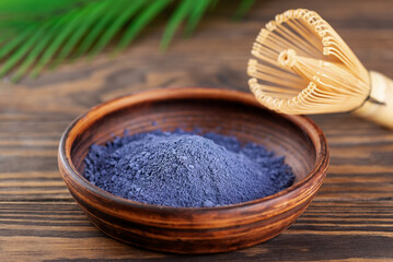Blue matcha powder from clitoria flowers in a bowl on a wooden table with chasen and palm branch on a background.