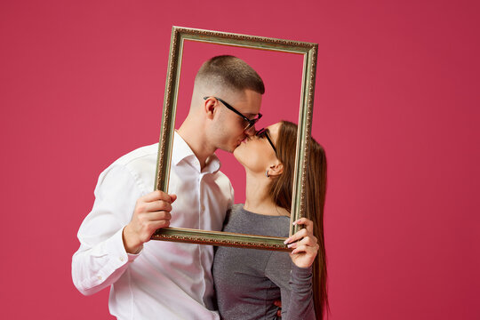 Portrait of happy young couple, man and woman, posing with picture frame, kissing against pink studio background. Concept of love, relationship, Valentine's Day, emotions, lifestyle