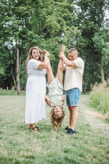 Obraz premium Mom and dad are standing in nature and holding their daughter's legs, which is hanging upside down.