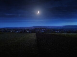 Crescent moon in a cloudy night sky. The moon is over the fields and the village in the distance. Beautiful night panorama.