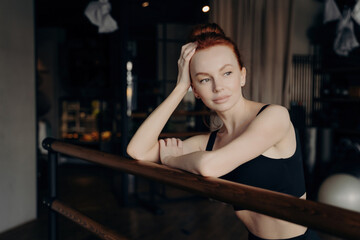 Fototapeta na wymiar Young attractive woman with red hair stands leaning on ballet barre and resting after workout