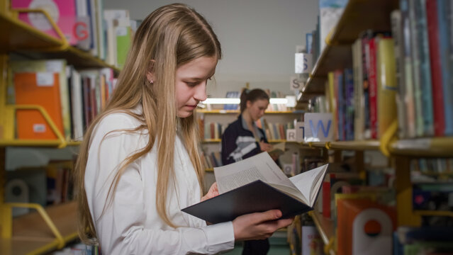 Curious female high school student inside the library, randomly picking books from the bookshelves. Education and knowledge concept.