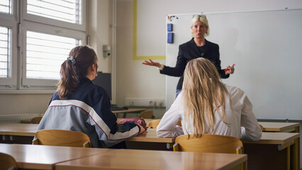 Rear view of two female students during a lecture in the classroom, looking at female teacher teaching and writing on a blackboard