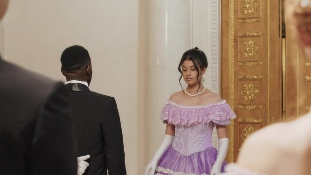 Over shoulder slowmo of Black man in elegant tuxedo suit nodding his head while inviting beautiful young Biracial debutante to dance at ball in pompous Regency style ballroom