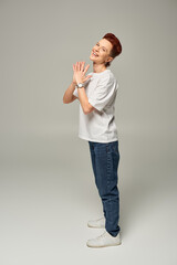Fototapeta na wymiar cheerful redhead bigender person in white t-shirt and jeans laughing and looking at camera on grey