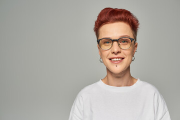 portrait of happy redhead queer person in white t-shirt and eyeglasses looking at camera on grey