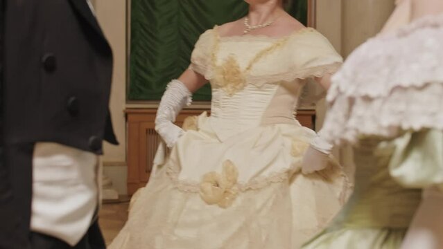 Tilting up shot of mid aged Caucasian lady holding poof skirt of her festive Regency dress while dancing on parquet and speaking to guests at ball ceremony