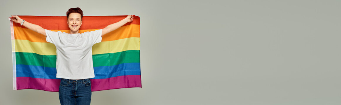 cheerful redhead bigender person in white t-shirt standing with LGBT flag on grey backdrop, banner