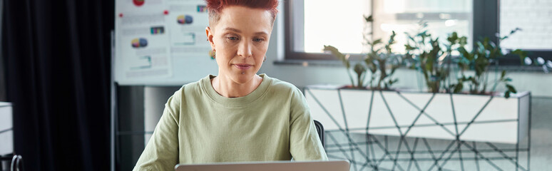 stylish ambitious queer person with short hair working at laptop in modern office, horizontal banner