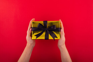 First person top view photo of hands holding golden giftbox with black ribbon bow over shiny golden sequins on isolated red background.