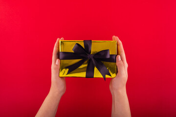 First person top view photo of hands holding golden giftbox with black ribbon bow over shiny golden sequins on isolated red background.